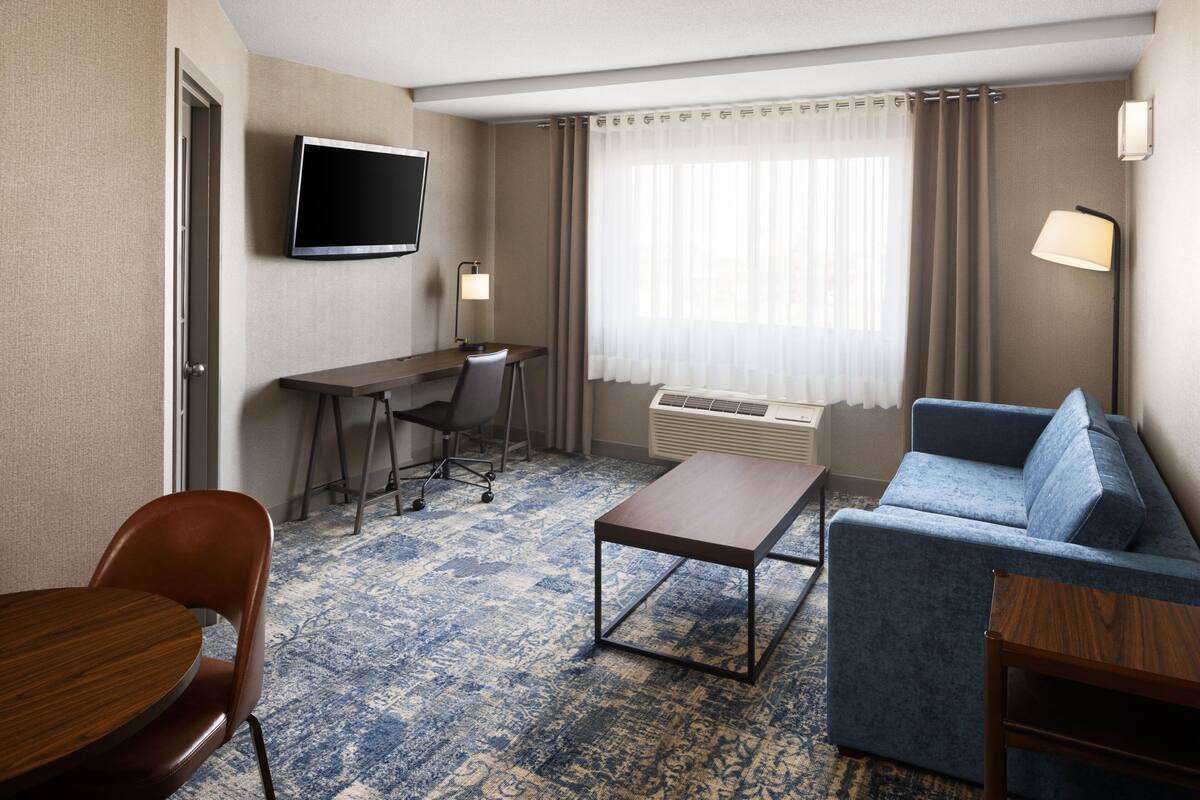 A hotel room with a desk, a blue sofa, a coffee table, a TV, and a window with curtains.