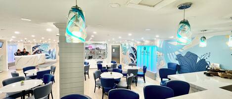 Un restaurante moderno con mesas redondas y sillas azules, que presenta una barra central de mármol y decoración mural.