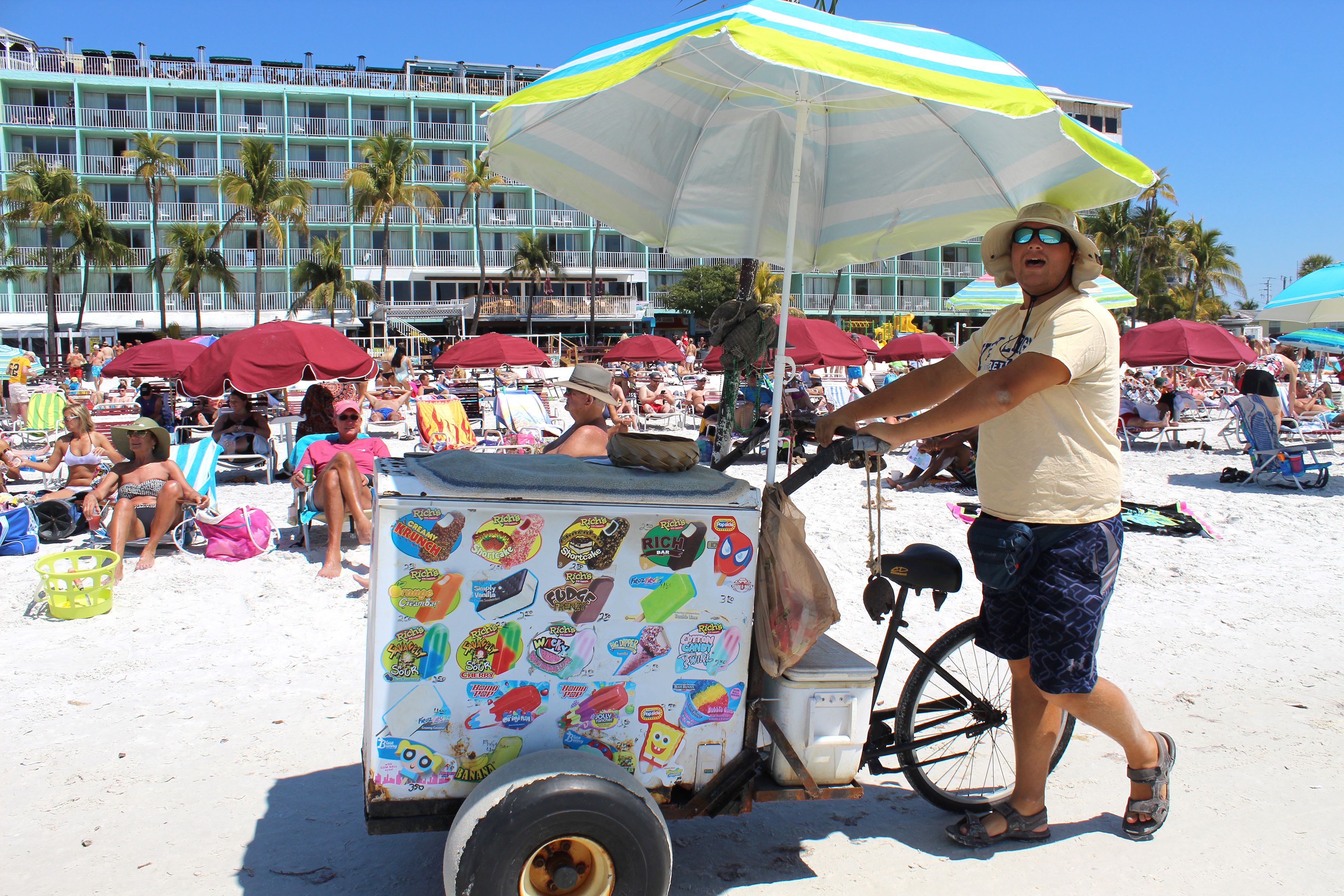 A beach scene with a man pushing a cart covered in colorful stickers, palm trees, and beach umbrellas in the background.