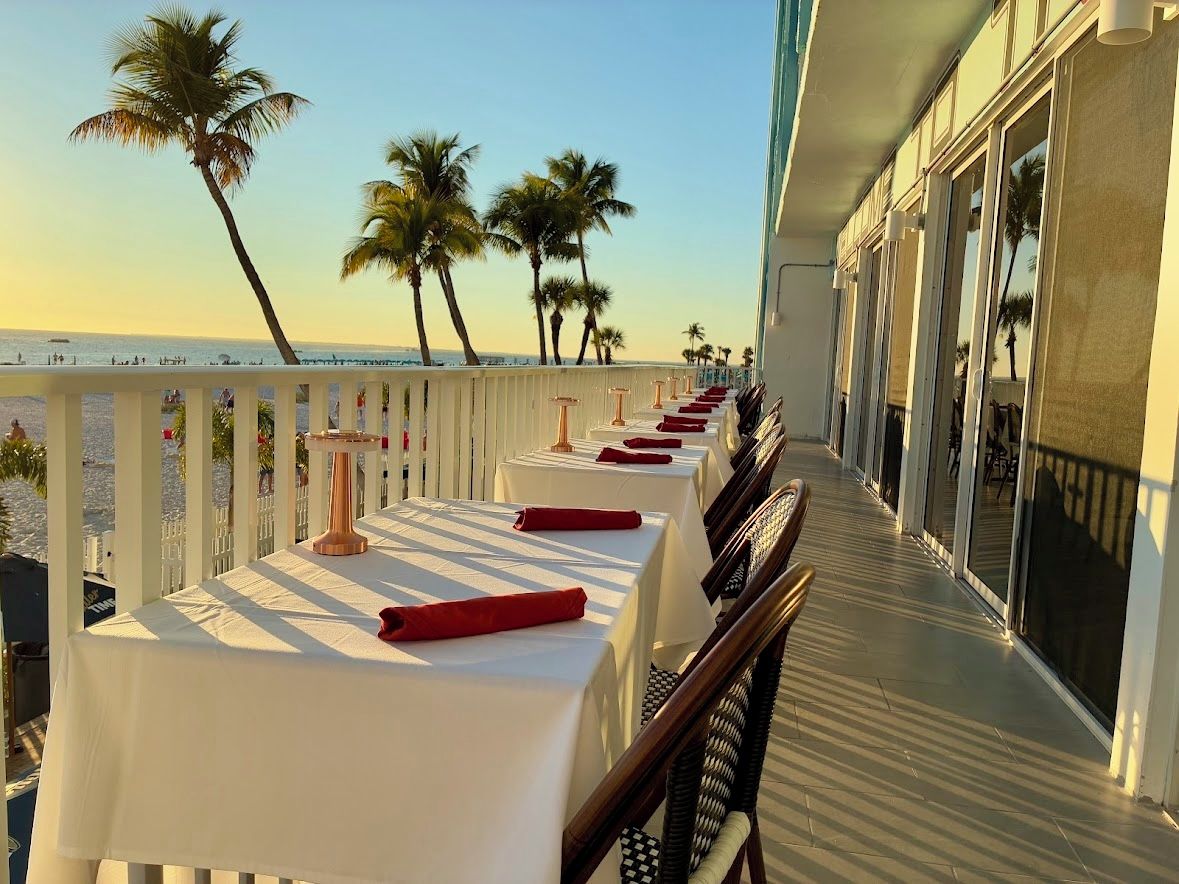 A balcony with tables set for dining, overlooking a beach with palm trees and the ocean.