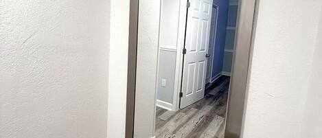 Un pasillo con un espejo grande en la pared, suelo de madera y una puerta blanca al final.
