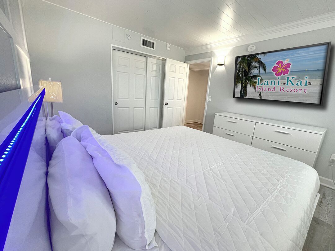 A bedroom with a bed, white walls, a TV displaying 'Lani-Kai Island Resort', and a door leading to another room.