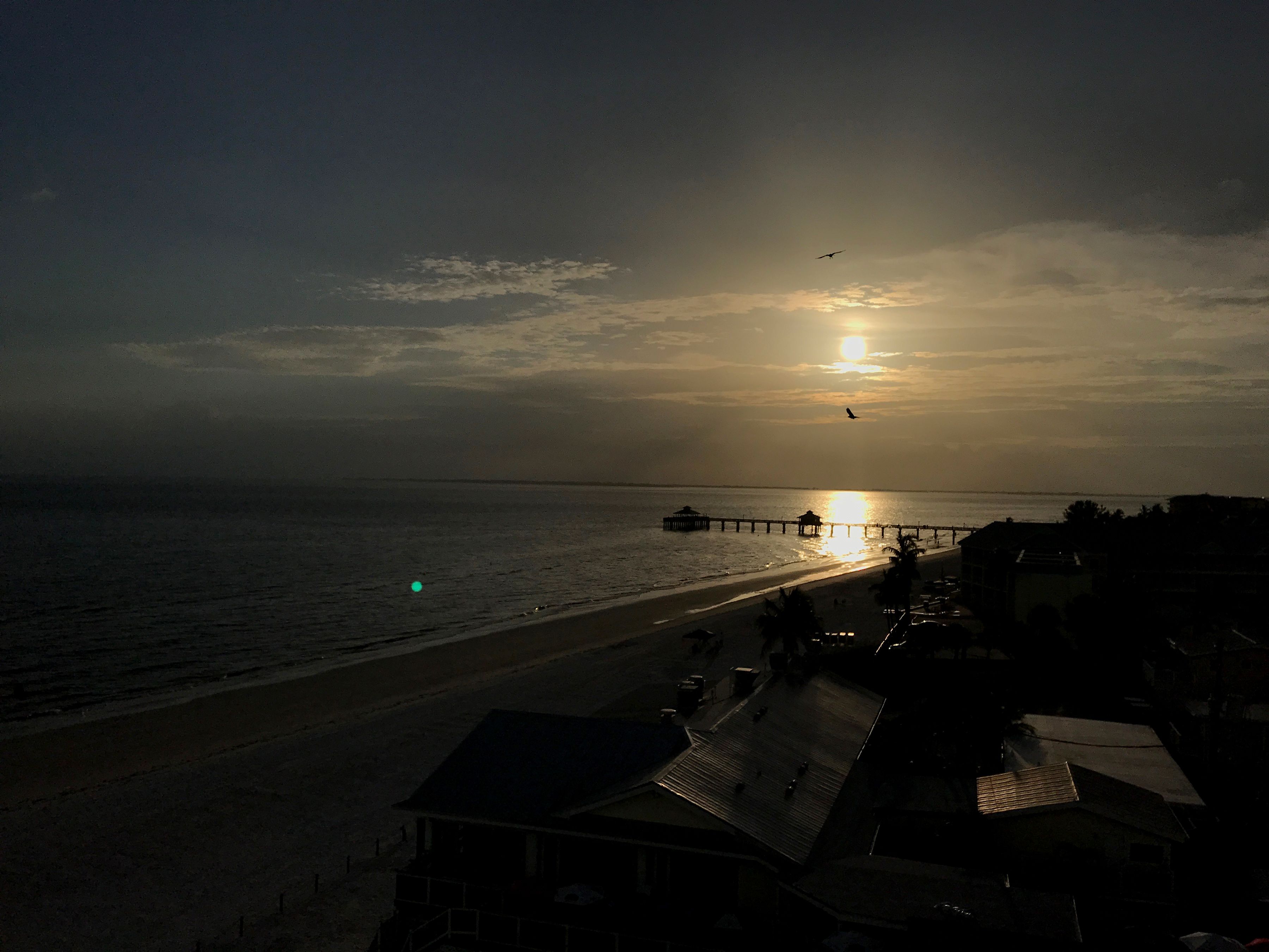 Blick auf einen Strand mit einem Steg, der sich ins Wasser erstreckt, einem Sonnenuntergang im Hintergrund und einigen Gebäuden im Vordergrund.