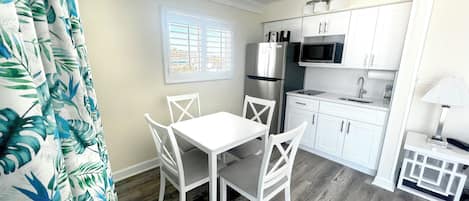 A compact kitchen with white cabinetry, a stainless steel refrigerator, a microwave, and a sink. There is a small dining table with four chairs.
