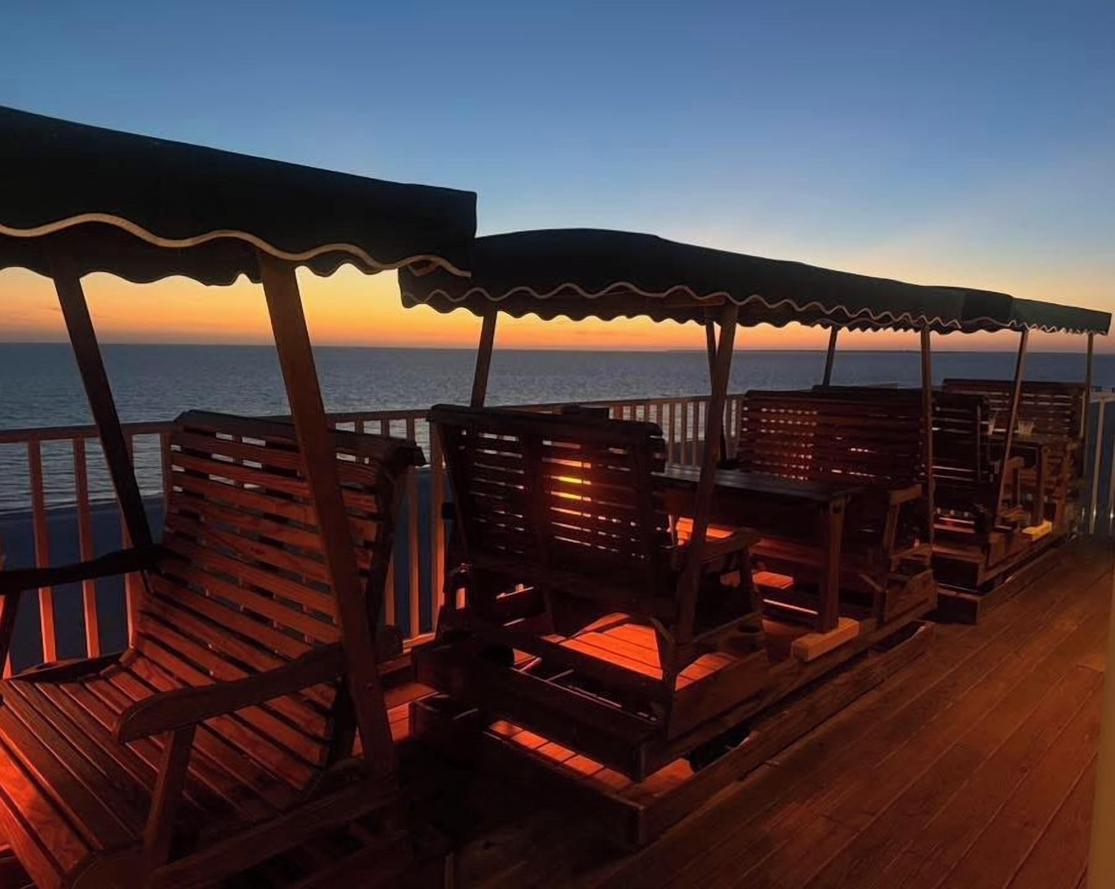 Une terrasse en bois, dotée de bancs et d’une rambarde, qui offre une vue sur la mer au coucher du soleil.