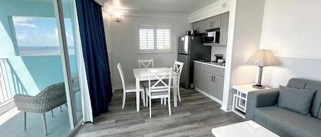 Una sala de estar moderna con un sofá gris, una mesa de centro blanca y un comedor con sillas y una mesa blancos. Se observa una cocina con armarios blancos y un refrigerador. Un ventanal con cortinas azules ofrece vistas al exterior.