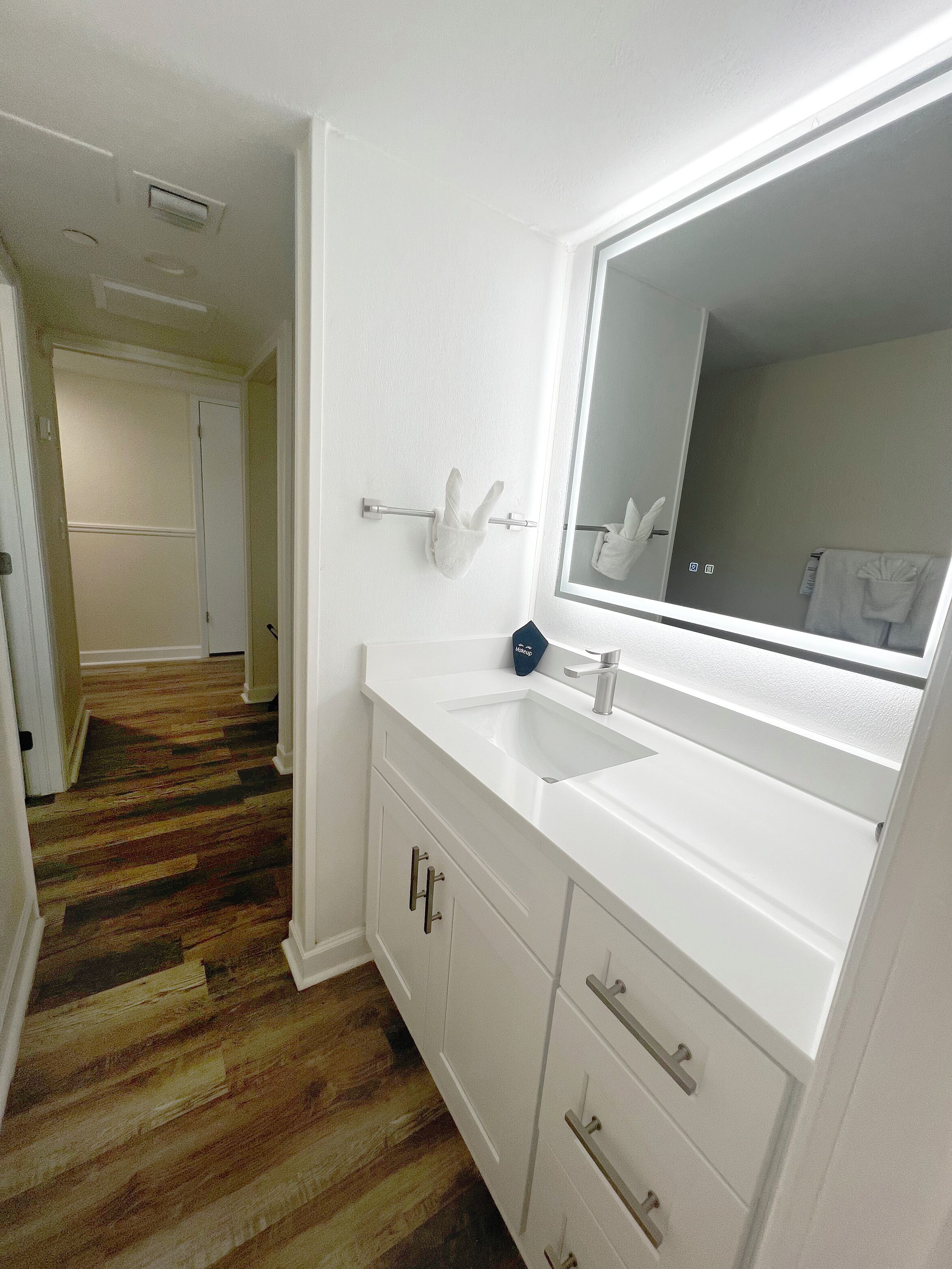 A bathroom with a white vanity, a large mirror, and a wooden floor.