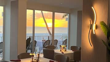 Ein Restaurant mit Tischen und Stühlen und Blick auf den Ozean, während die Sonne untergeht.