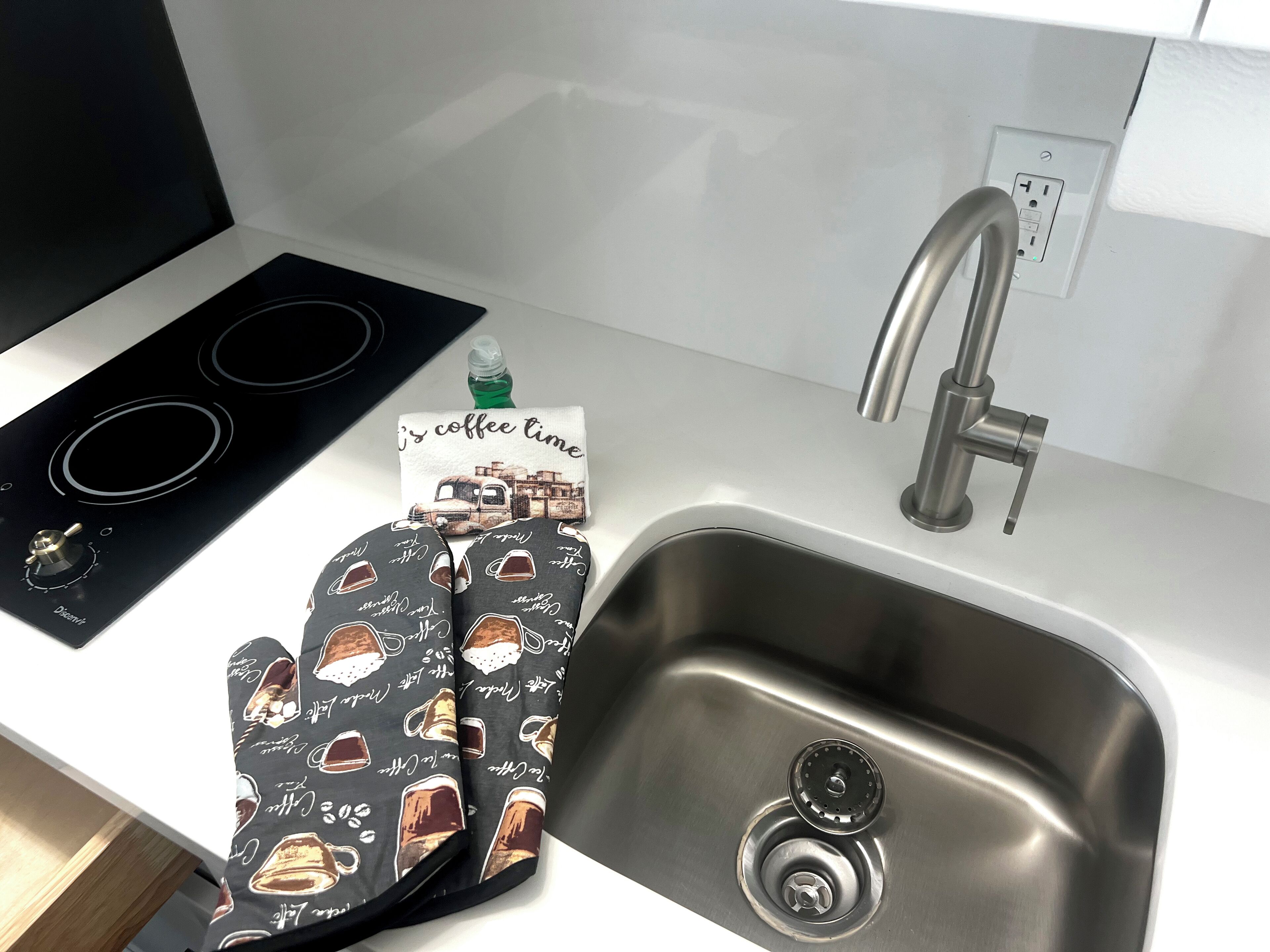 Une cuisine équipée d’un évier en acier inoxydable, d’un robinet, d’une plaque de cuisson noire et d’un torchon à motifs de café.