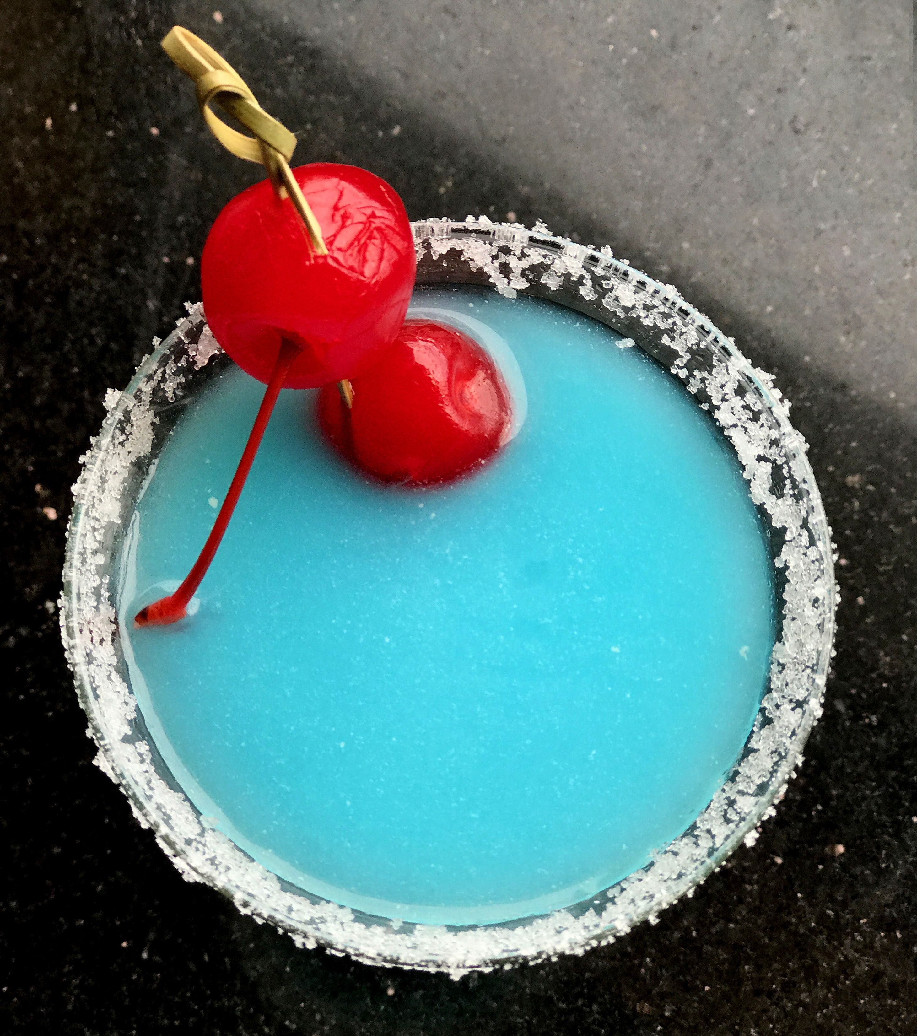 Blauer Drink in einem Glas mit einer roten Kirsche am Spieß, umrandet von einer Salzkruste.