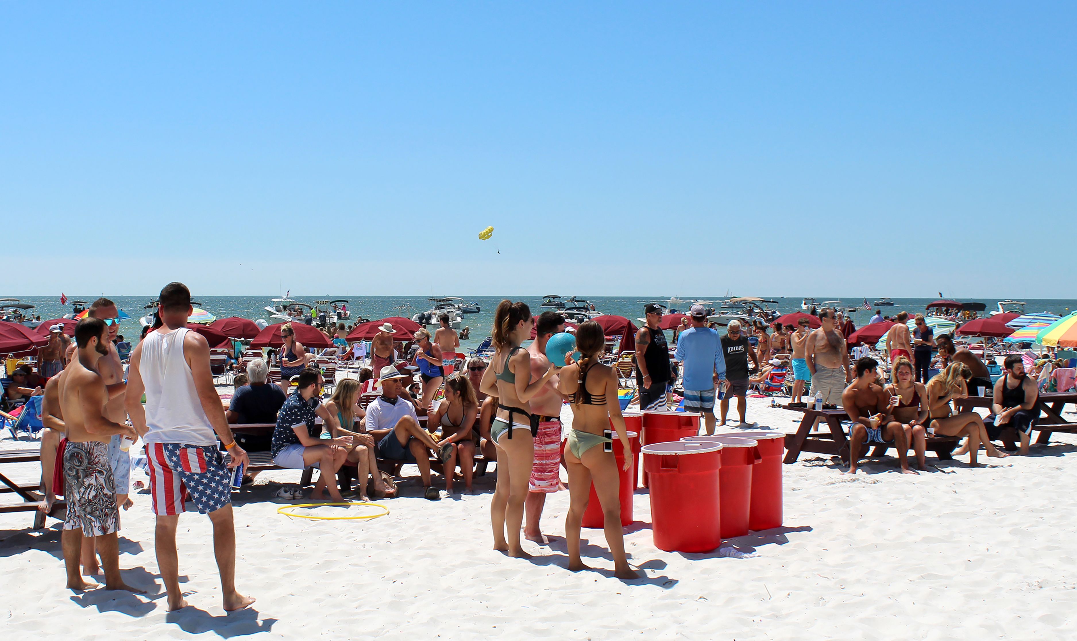 A crowded beach with people, umbrellas, and boats in the background.