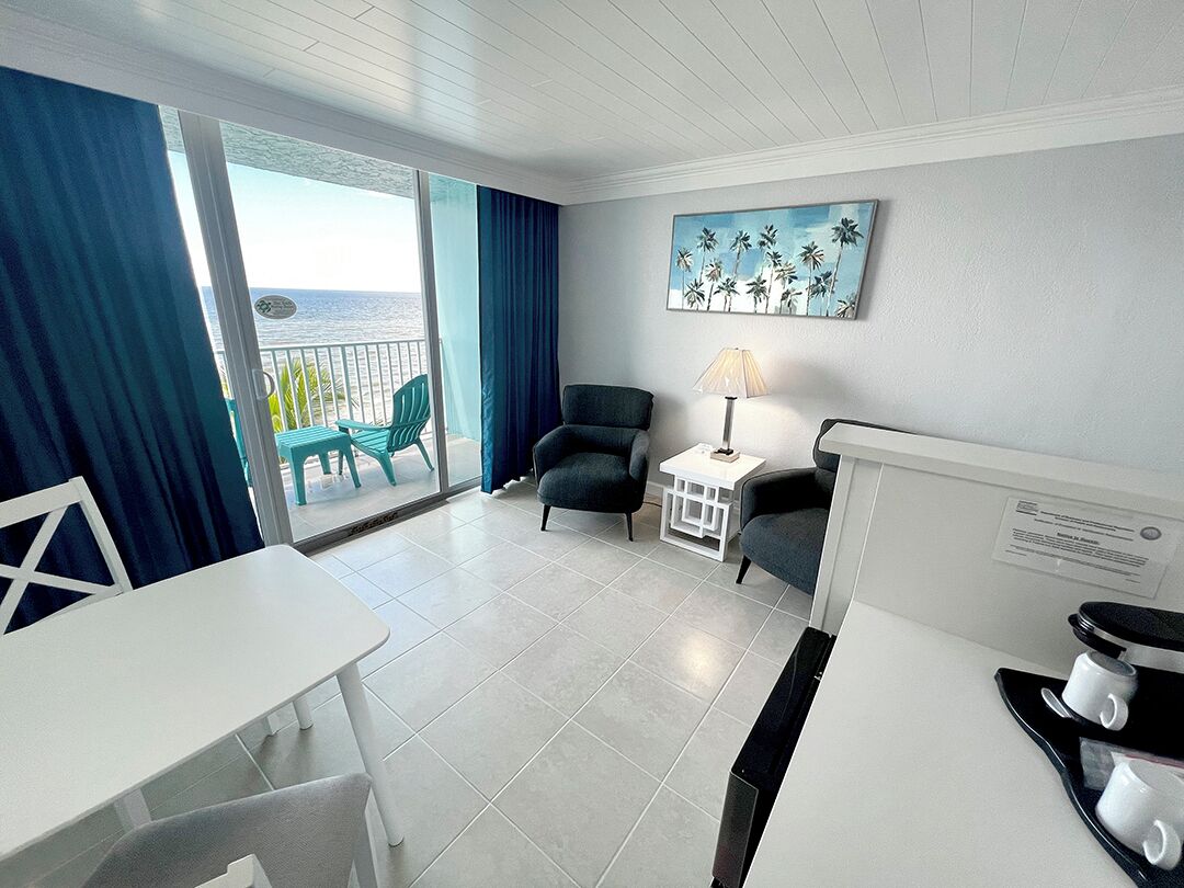 A hotel room with a balcony, a white table, two chairs, a small white side table with a lamp, and a painting of palm trees.