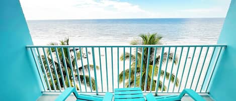 A balcony with two blue lounge chairs and a small table, overlooking a beach with palm trees and the ocean.