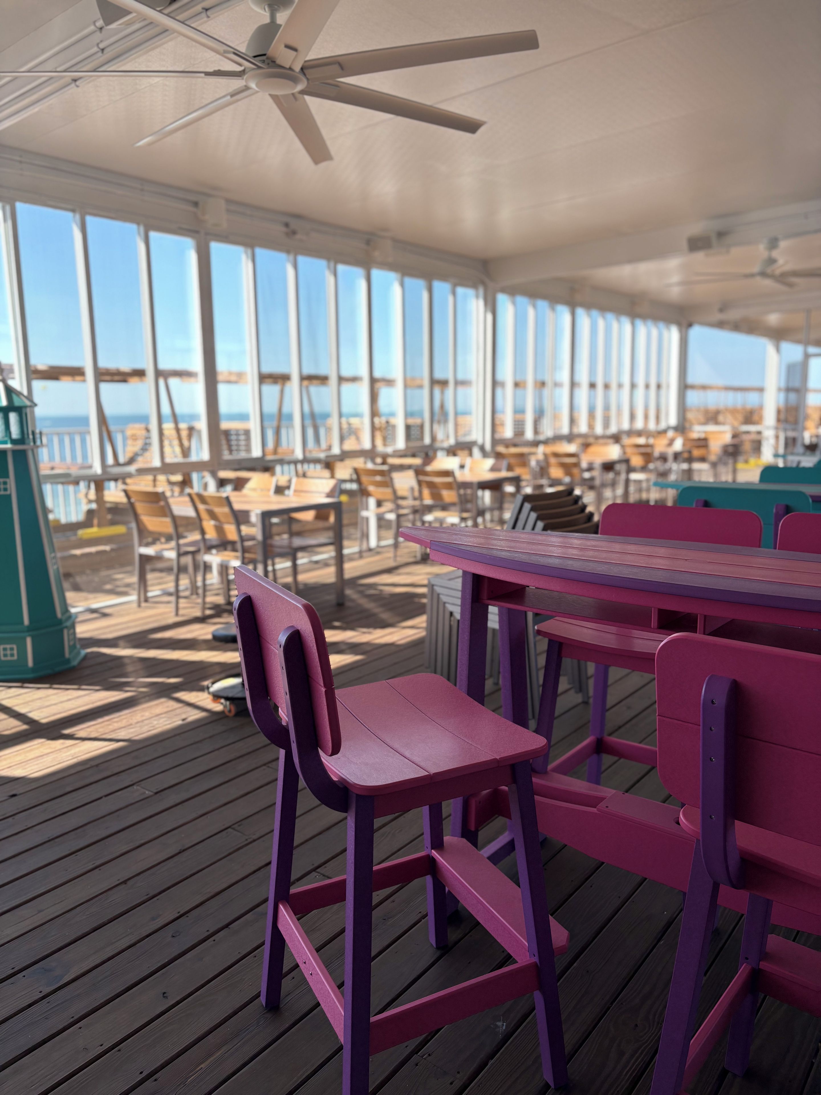A covered outdoor seating area with tables and chairs, large windows, and a view of the ocean.