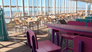 A covered outdoor seating area with tables and chairs, large windows, and a view of the ocean.