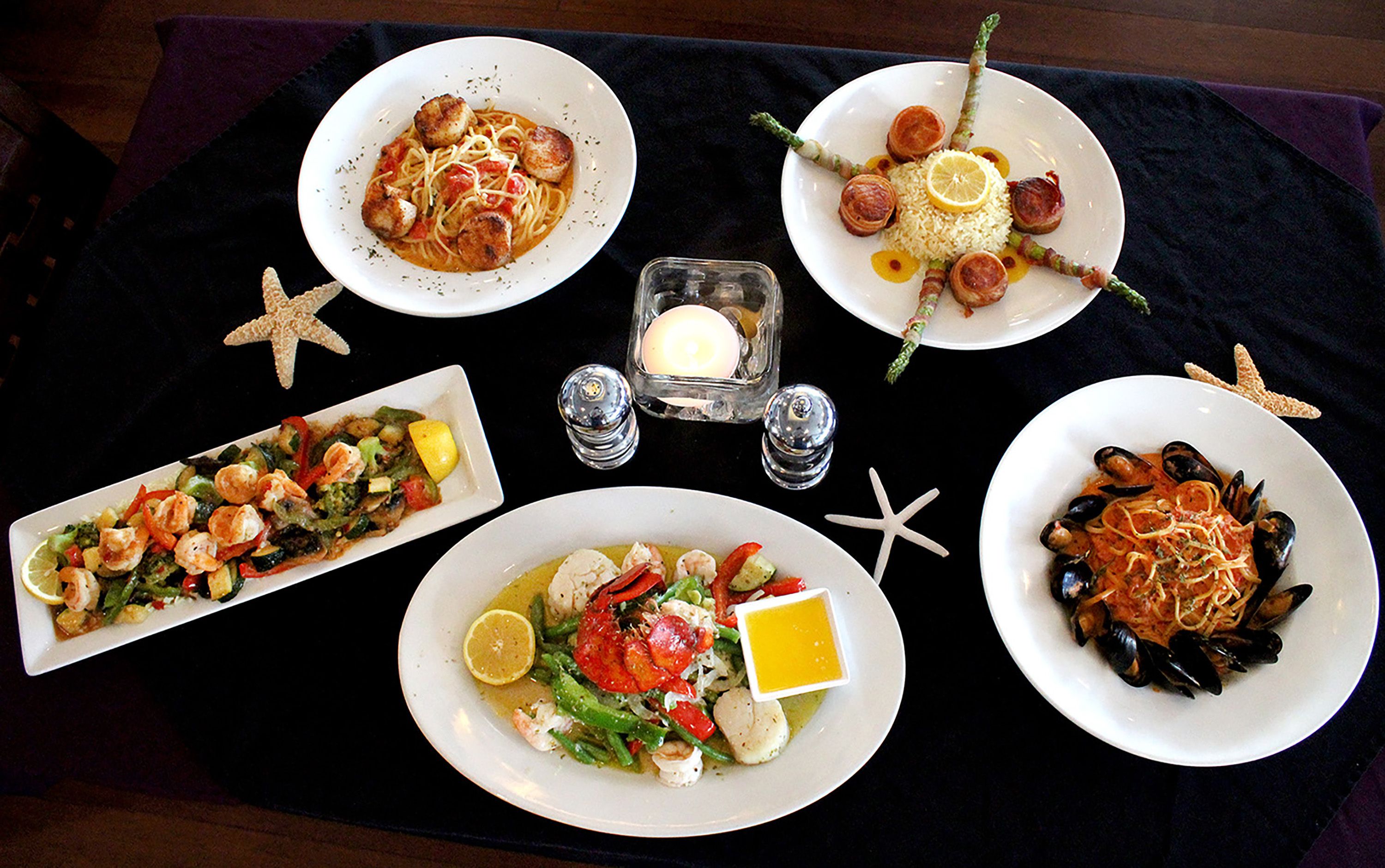 A table set with various seafood dishes, including pasta, shrimp, and mussels, arranged on a black tablecloth.