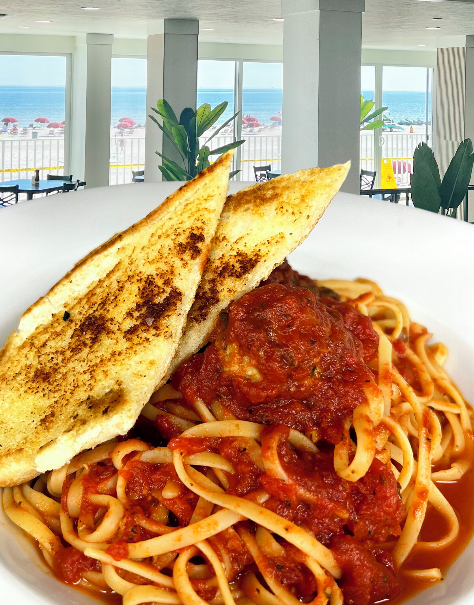 Un plato de espagueti con salsa de tomate y pan a la parrilla, frente a una vista de la playa y sombrillas.