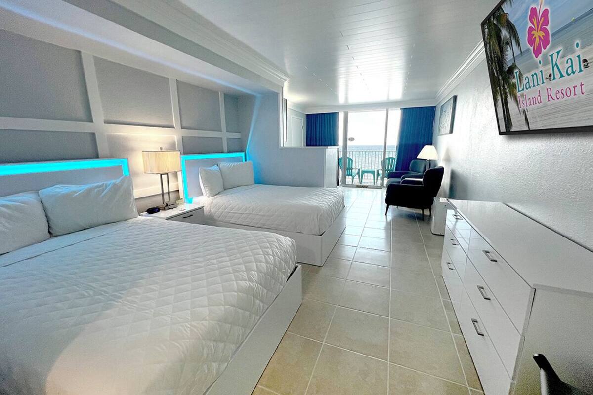 A hotel room with two beds, a desk, a chair, and a balcony with a view of the sea.