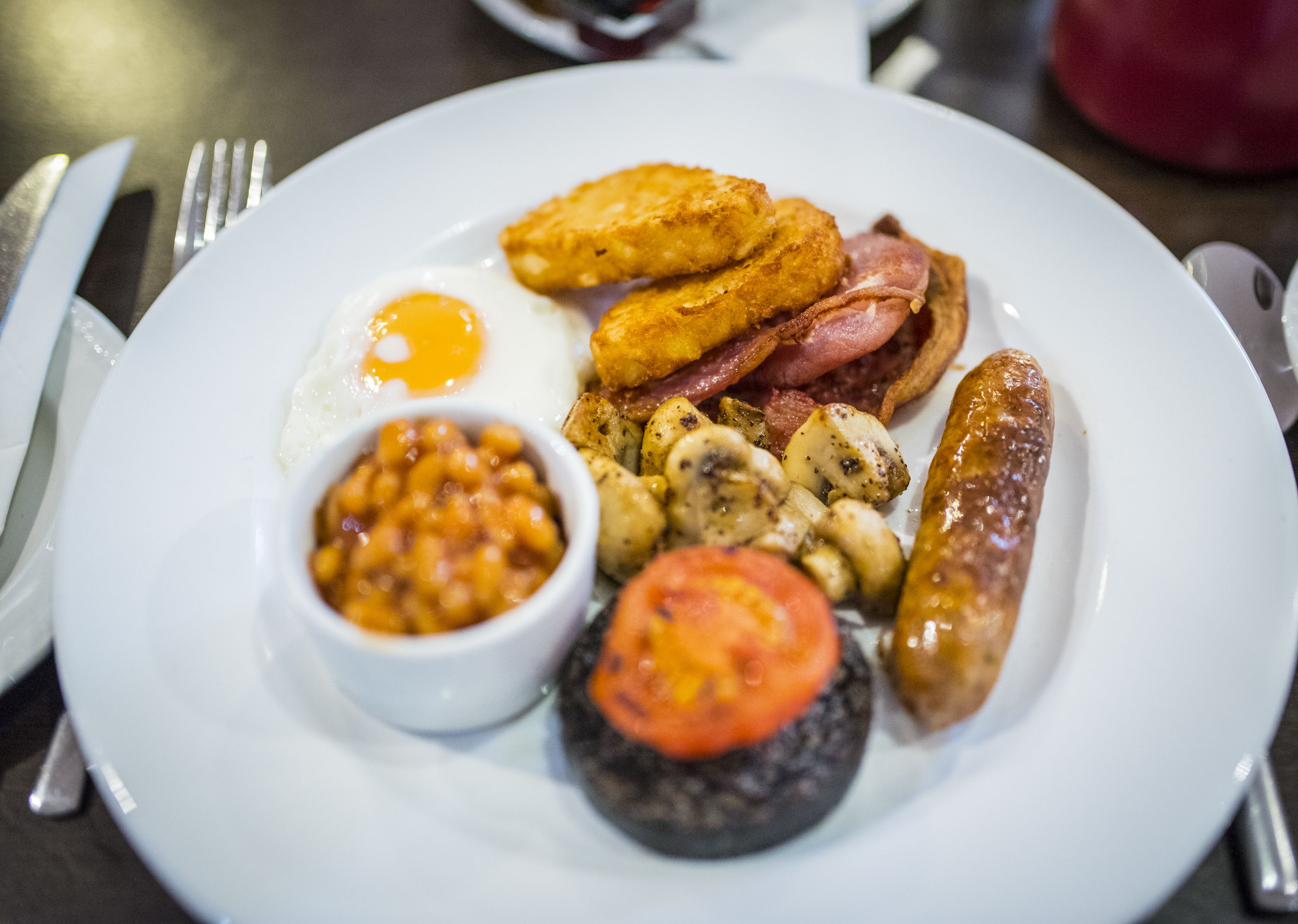 A plate of breakfast food including eggs, bacon, sausages, mushrooms, baked beans, and a tomato.