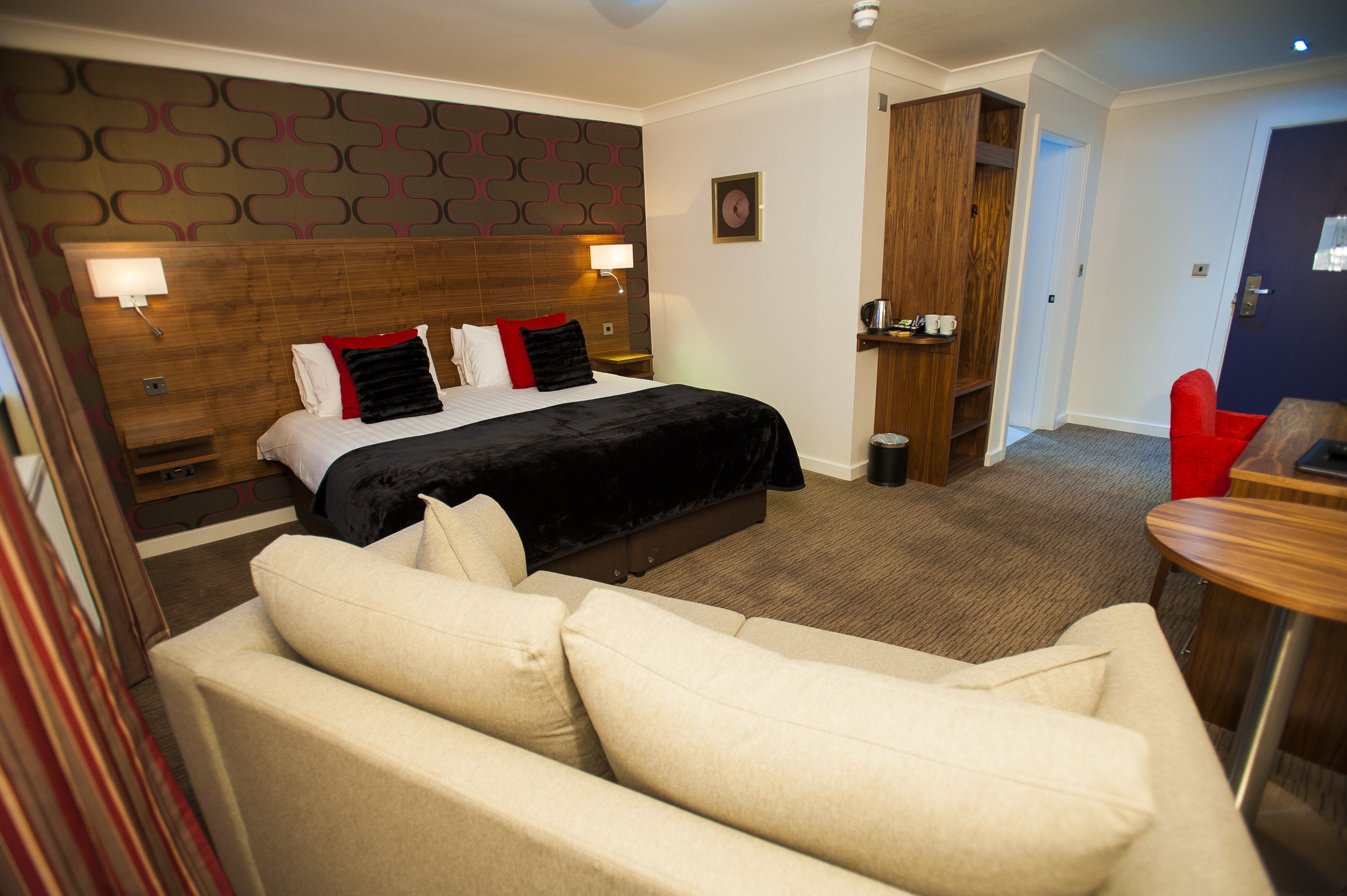 A hotel room with a bed, a sofa, a wooden headboard, a wall clock, and a door leading to another room.