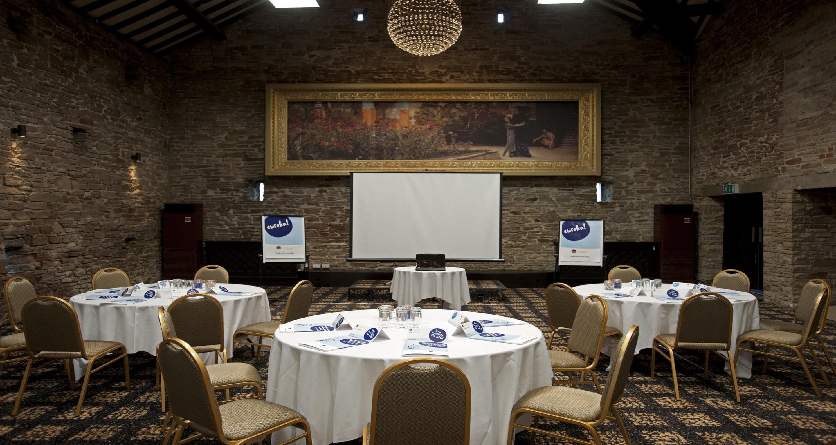 Un salón de conferencias con mesas redondas, sillas, una pantalla de proyector y un cuadro grande en la pared.