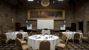 Un salón de conferencias con mesas redondas, sillas, una pantalla de proyector y un cuadro grande en la pared.