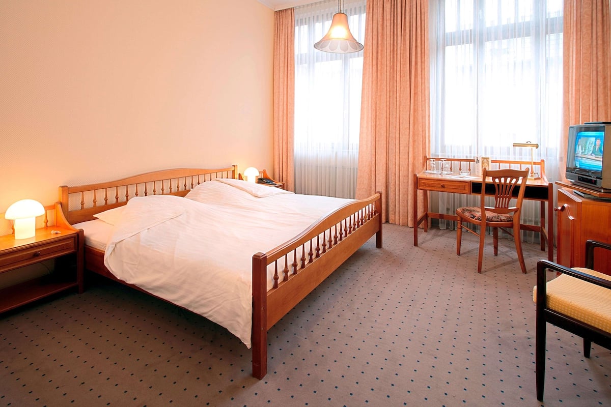 A hotel room with a wooden bed, a desk with a chair, a television, and a window with curtains.