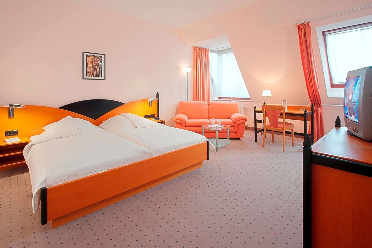 A hotel room with a double bed, a sofa, a desk, a chair, a TV, and a window with curtains.