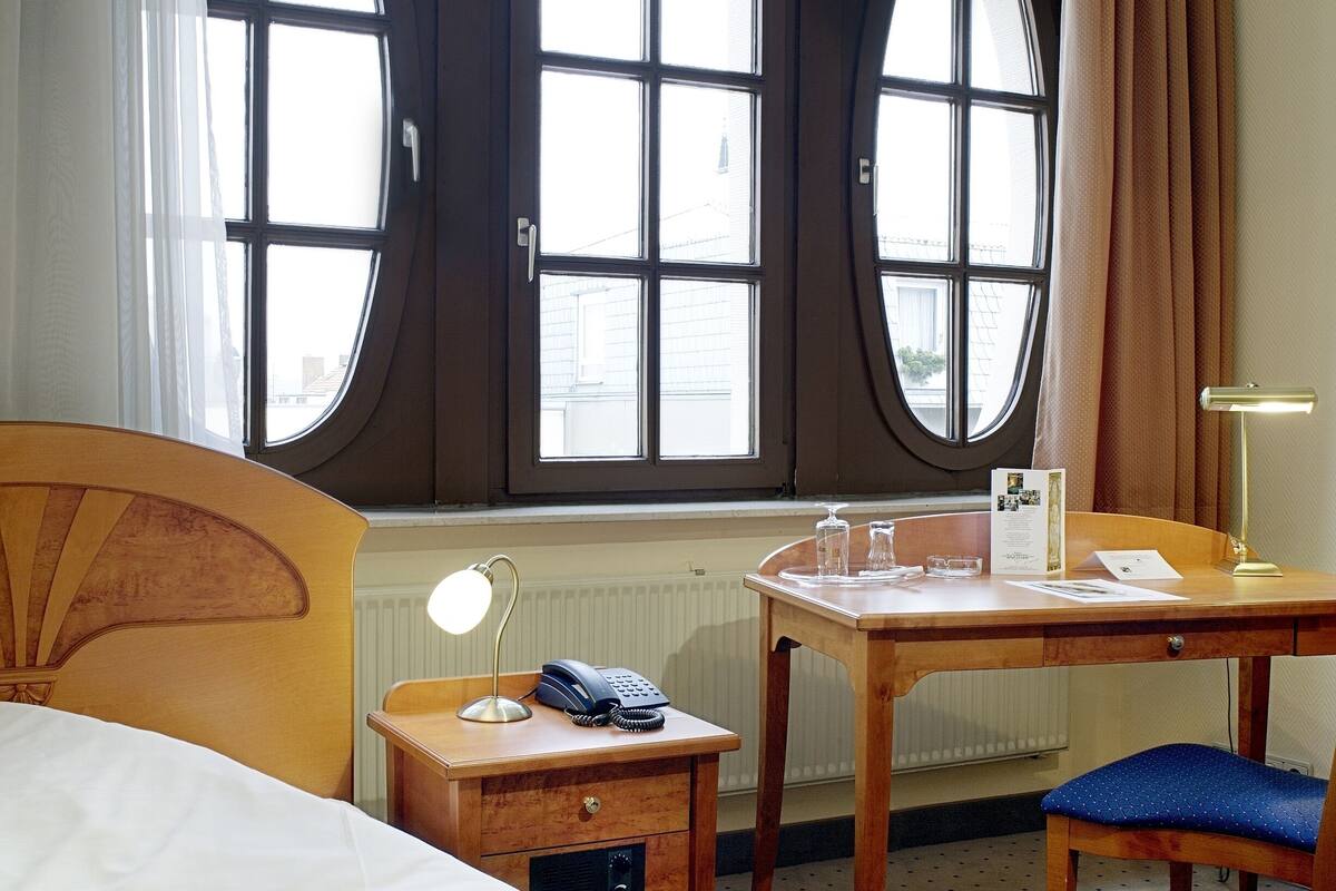 A hotel room with a bed, a desk with a telephone, a chair, and a window with curtains.