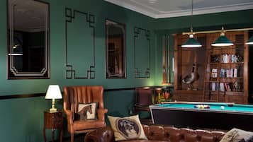 Una habitación con mesa de billar, sillones de cuero, una estantería y un espejo grande.