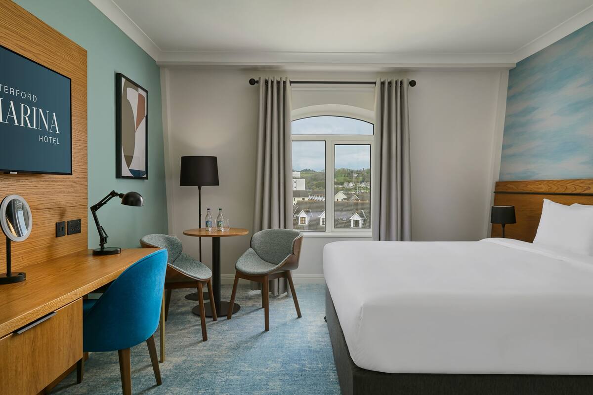 Une chambre d’hôtel avec un lit, un bureau, des chaises, une petite table avec des bouteilles, une lampe et une vue sur les bâtiments par la fenêtre.