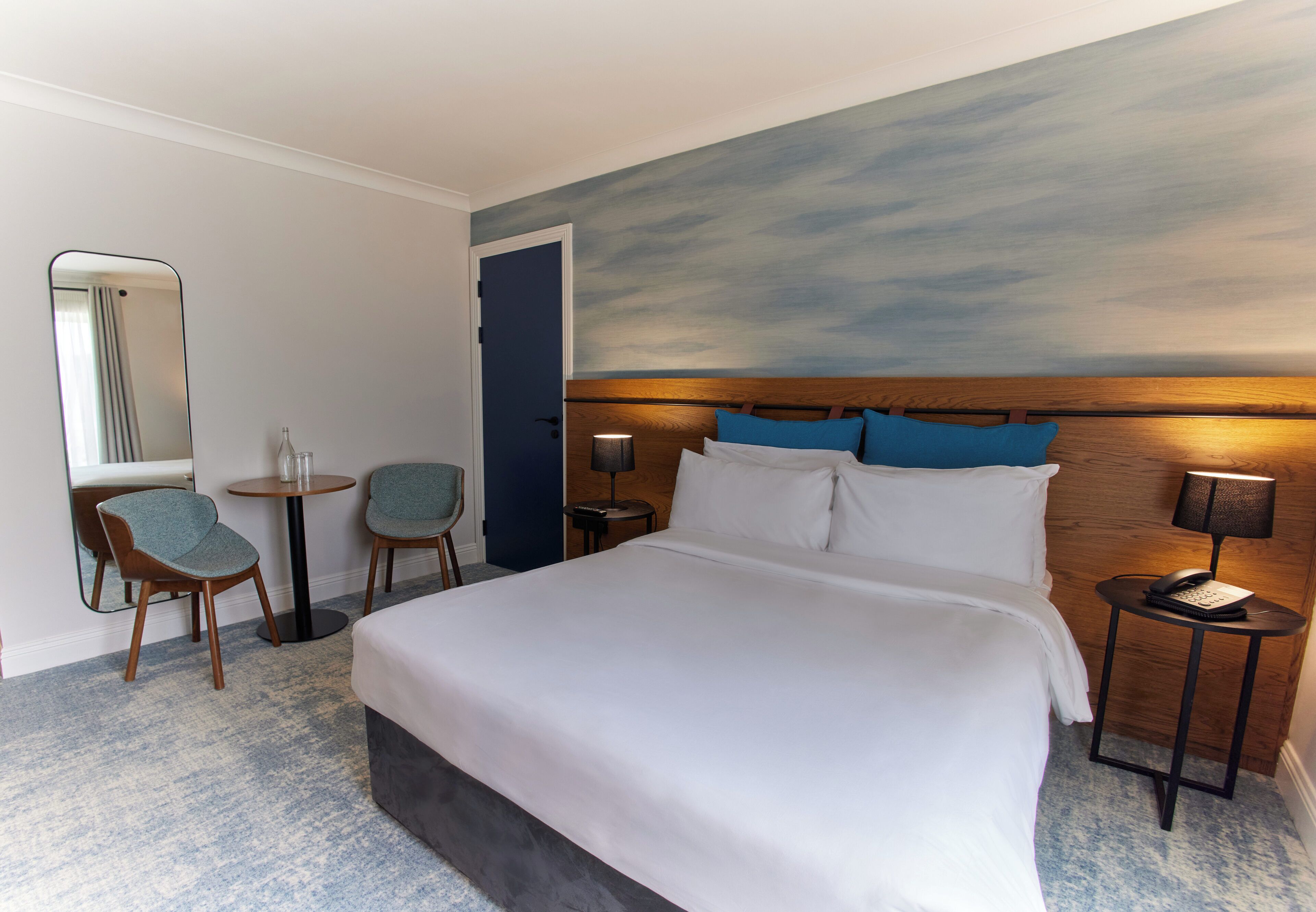 Una habitación de hotel con una cama grande, dos sillas, una mesita, una mesita de noche con lámpara y un espejo colgado en la pared.