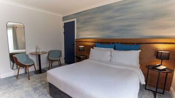 A hotel room with a large bed, two chairs, a small table, a nightstand with a lamp, and a wall-mounted mirror.
