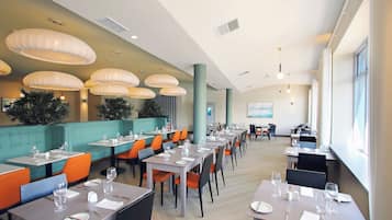Uma sala de jantar espaçosa com várias mesas e cadeiras, grandes luminárias pendentes e vista para a cidade através de amplas janelas.