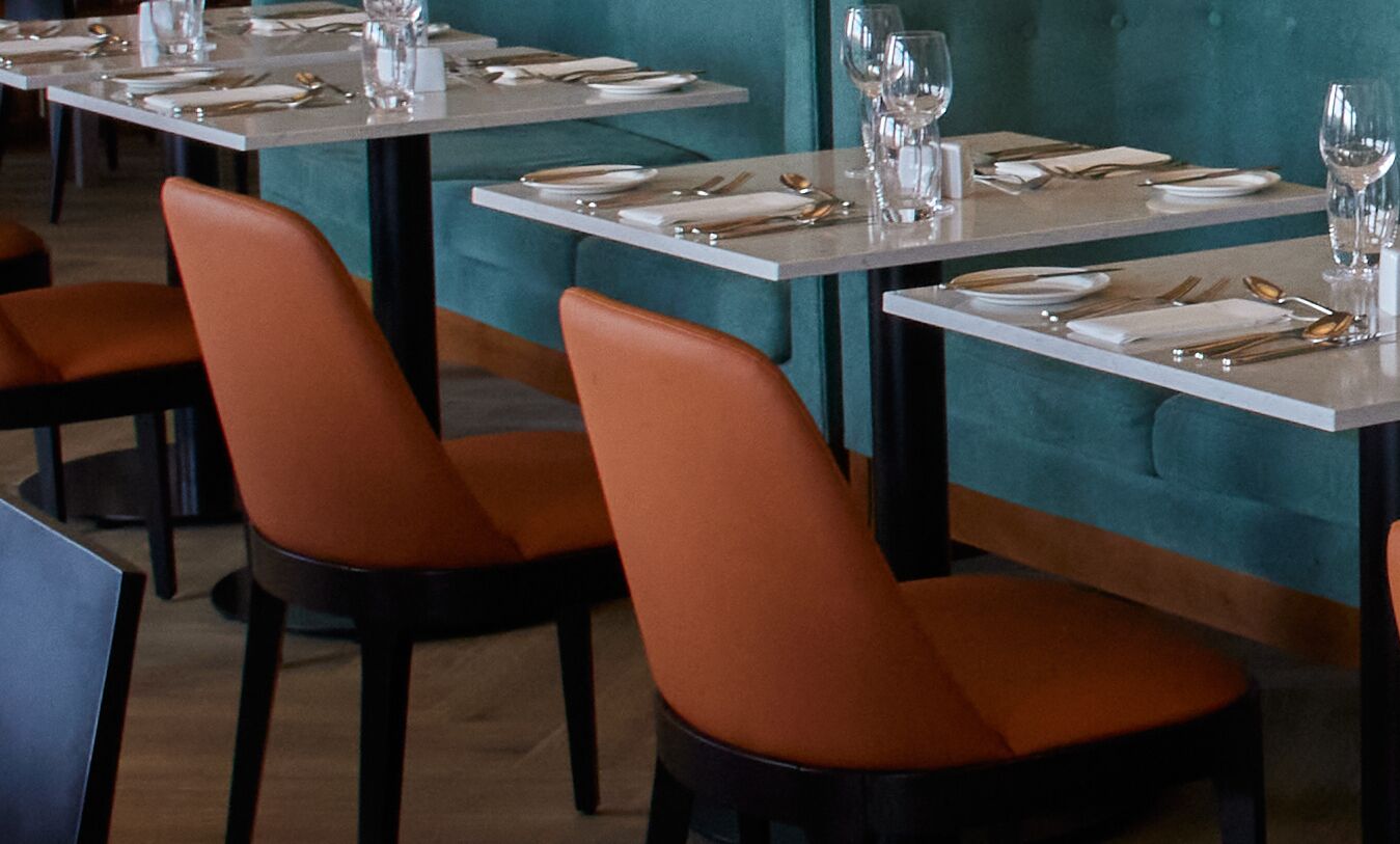 Un restaurante con mesas listas para comer, con sillas color naranja y tapicería verde azulado.