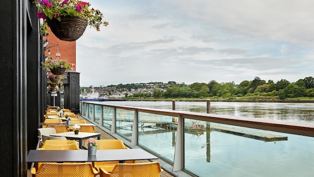 Een terras aan de rivier met gele rieten stoelen en tafels, uitzicht op de rivier, en bloembakken.