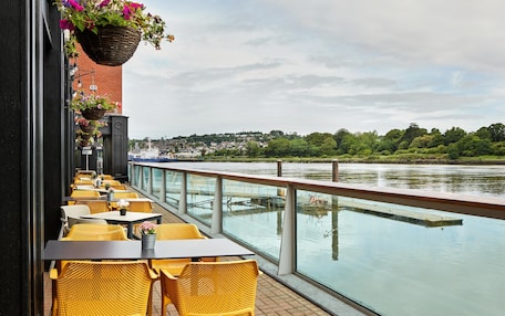 Terraza a la orilla del río con sillas y mesas de mimbre amarillo, vistas al río y macetas colgantes de flores.
