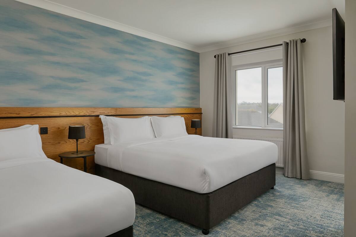 Une chambre d’hôtel avec deux lits, une tête de lit en bois, une grande fenêtre avec des rideaux et une moquette bleue à motifs.
