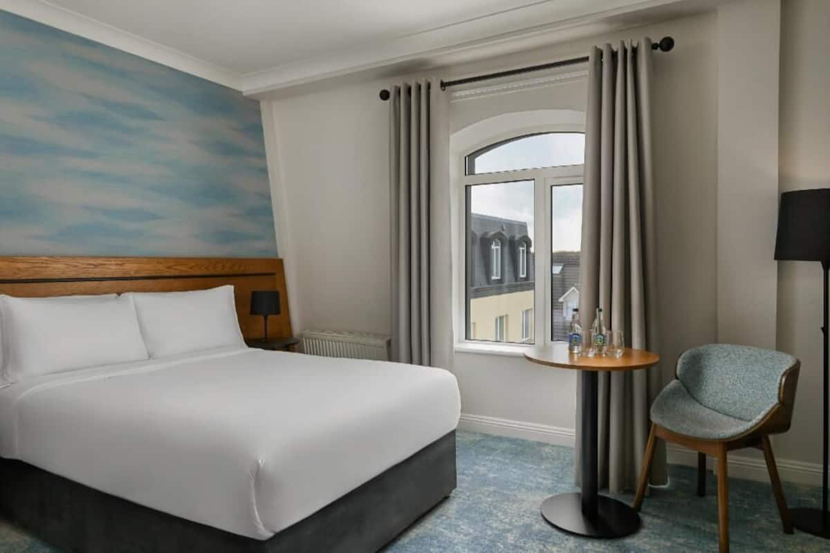 Ein Hotelzimmer mit einem Bett, einem runden Tisch mit Stuhl, einem Fenster mit Vorhängen und einer Wand mit Himmel-Fresko.