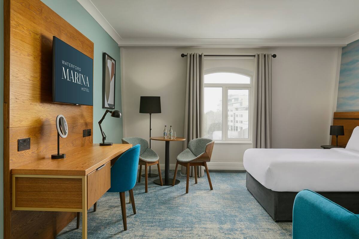 Ein Hotelzimmer mit einem Bett, einem Schreibtisch mit Stuhl, einem kleinen Tisch für zwei Personen, einem an der Wand befestigten Fernseher und einem Fenster mit Vorhängen.