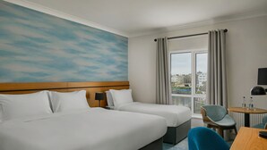 Hotelkamer met twee bedden, een klein tafeltje met stoel, een raam met gordijnen en uitzicht op de stad.