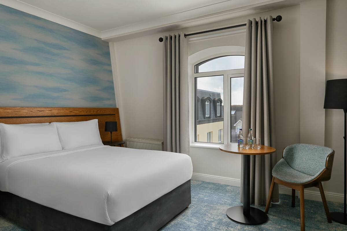 Une chambre d’hôtel avec un lit, une table ronde et une chaise, une fenêtre avec des rideaux et une vue sur les bâtiments environnants.