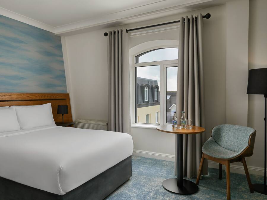 A hotel room with a bed, a round table with a chair, a window with curtains, and a view of buildings outside.