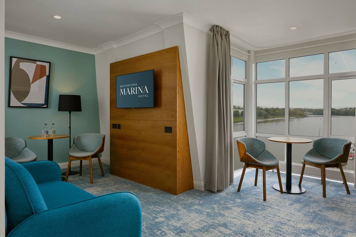 Ein Hotelzimmer mit großem Fenster, einer Wand mit Holzvertäfelung, einem Fernseher und einer Sitzecke mit Sesseln und einer Couch.