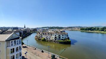 A riverside building with a prominent balcony, a pedestrian walkway, and a view of the river and distant cityscape.