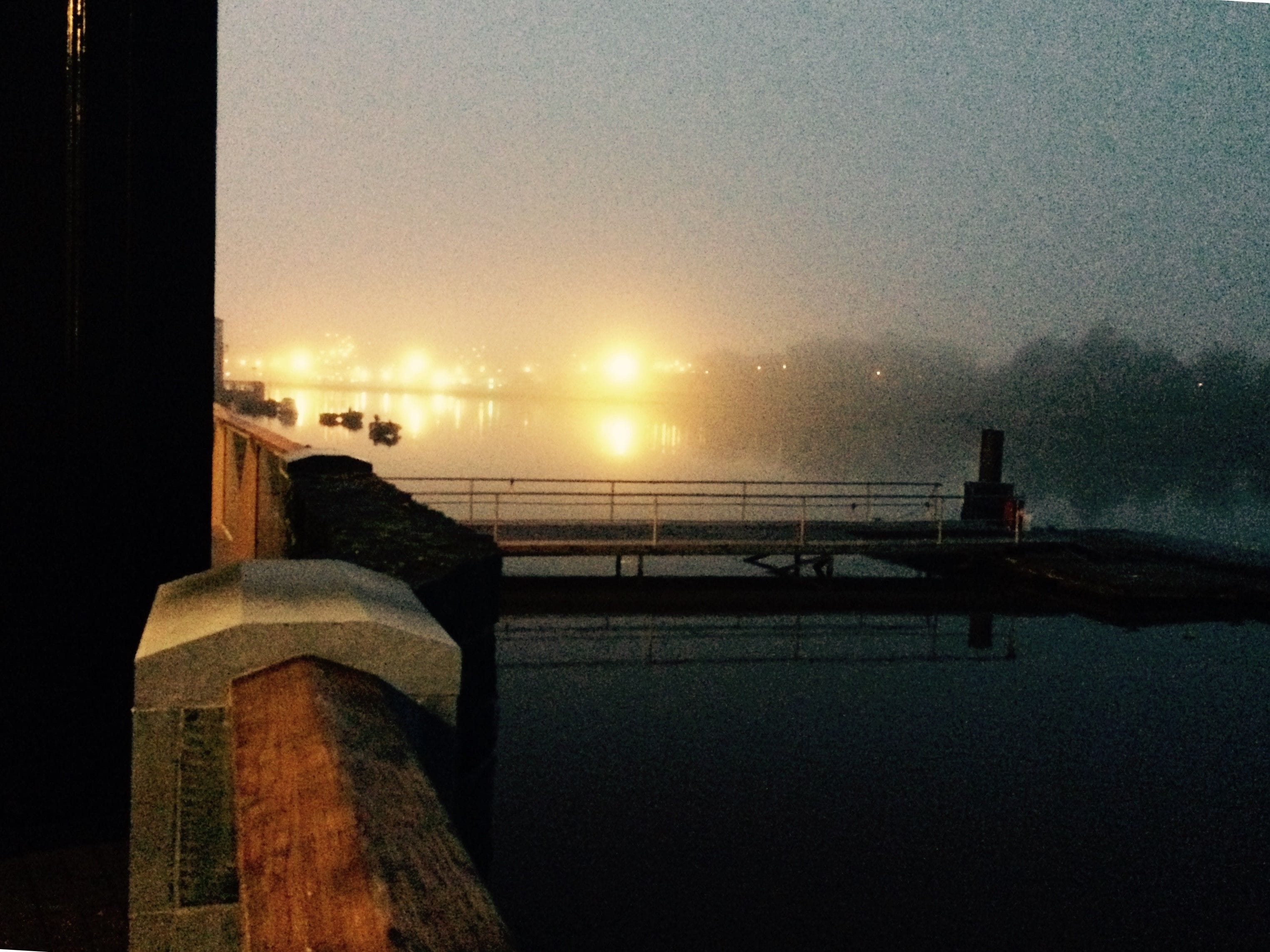 A waterfront view with a bridge, a railing, and distant lights reflecting on the water.