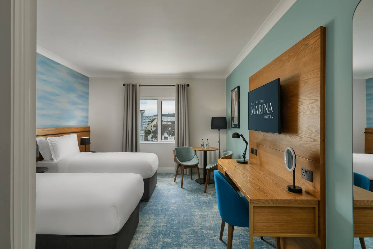 Une chambre d’hôtel avec deux lits, un bureau, une chaise, une table ronde et une vue sur la ville par la fenêtre.