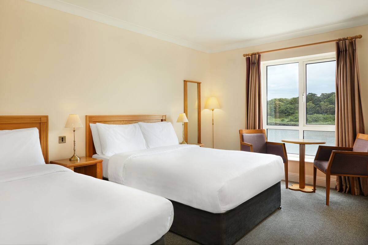 Ein Hotelzimmer mit zwei Betten, einem kleinen Tisch, zwei Stühlen, einem Fenster mit Vorhängen und Blick auf Grünflächen.
