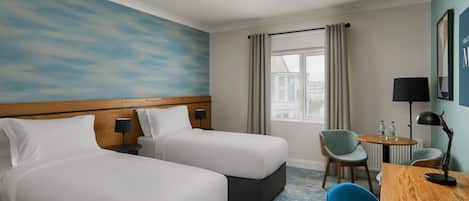 A hotel room with two beds, a round table with chairs, a lamp, and a view of the city through a window.