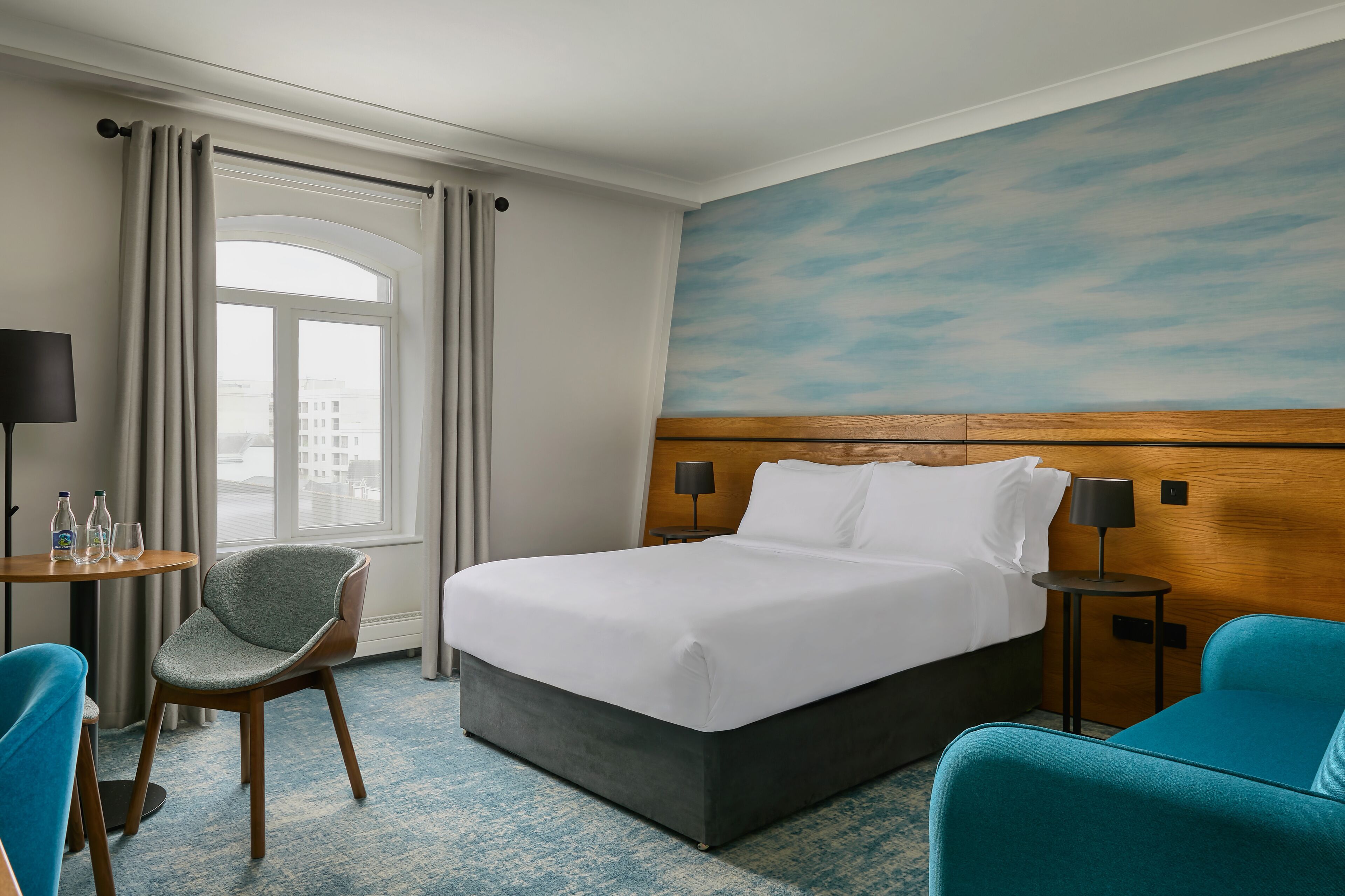 A hotel room with a bed, a chair, a small table with a lamp, and a view of the city through a window.